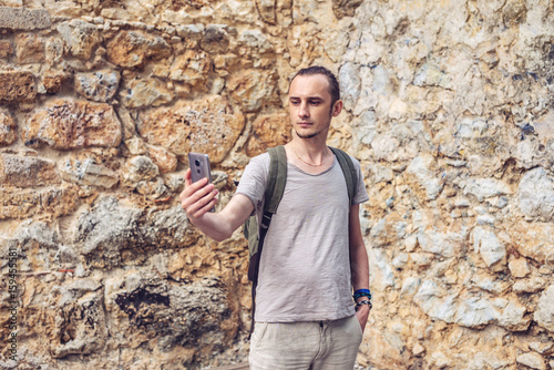 Man traveler with backpack makes a photo on his smartphone on the background of a stone wall