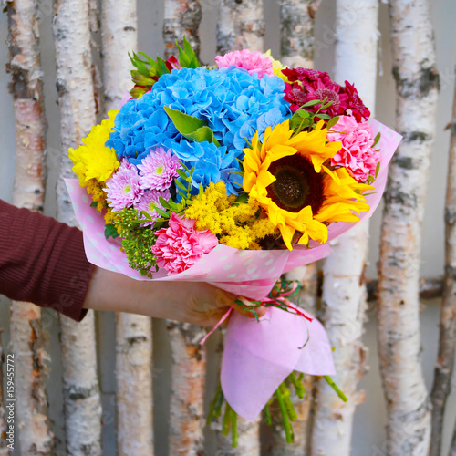 Fototapeta Naklejka Na Ścianę i Meble -  The florist holds in hands a beautiful bright summer bouquet of flowers on the background of wooden trunks. Blue hydrangea, sunflower, pink carnation, solidago