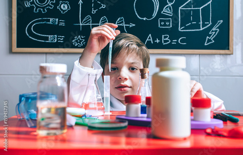 Portrait of little boy scientist extracting liquid from test tube behind of table with chemical toys