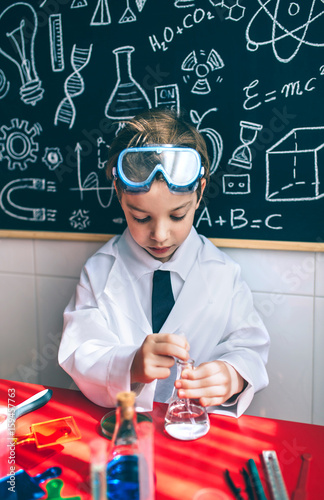Little boy scientist extracting liquid from flask against of chalkboard with drawings
