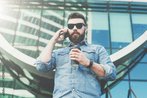 Young bearded hipster businessman in sunglasses and stands outdoor,drinking coffee and talks on his cell phone.