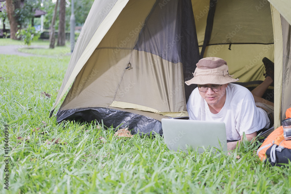 young man tourist lying and using laptop in tent at camping site in forest. Outdoor activity in summer. Adventure traveling in national park.