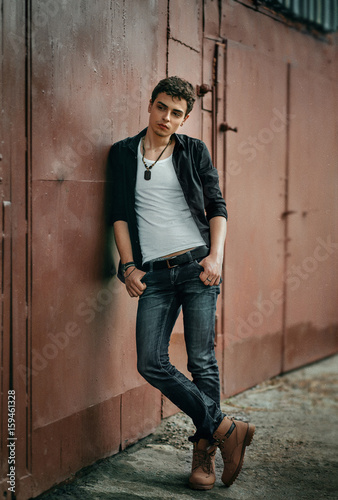 The young guy is standing near the garage. Poses for men's photo sessions. Beautiful appearance, clothes in rock style.