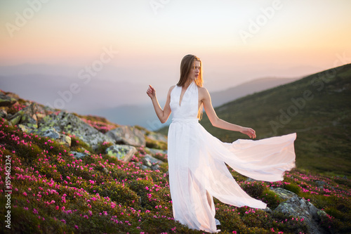 The girl in white dress with long flying train stands on the top of the rock. Woman in white dress walking in the mountains. Majestic flowers glowing by sunlight