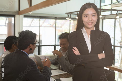asian businesswoman with folded hands smiling at camera. confident young woman wearing black suit with crossed arms standing at office with businessman have a meeting behind her.
