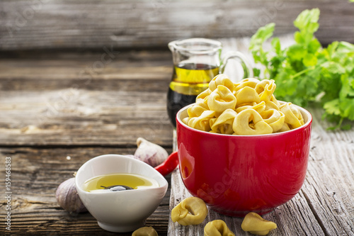 Homemade comfort food: Italian tortellini in a red ceramic bowl on a rustic wooden background with olive oil, black olives, herbs, garlic, balsamic sauce for cooking various dishes. Selective focus.