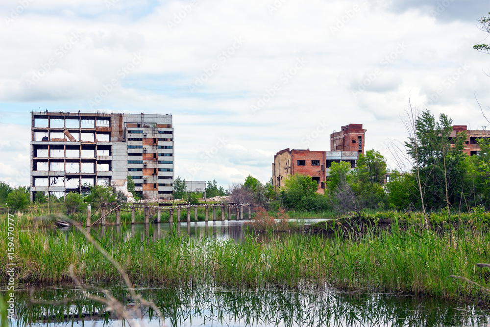 The abandoned building is located among the trees. There is a pond in front of the building. This is a former factory or office