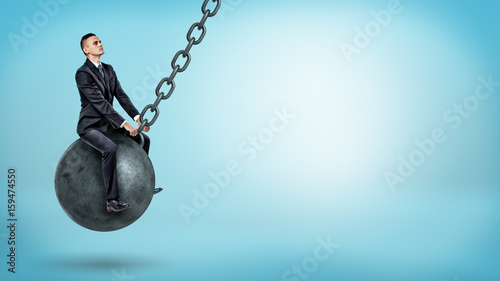 A businessman swinging on a large wrecking ball and looking up on blue background.