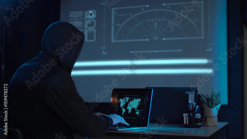 Man in the hood from his back sitting and working at laptop as hacker, with world map on wall in background. Then he take off usb flash disk and run away. Perhaps he has stolen information or has