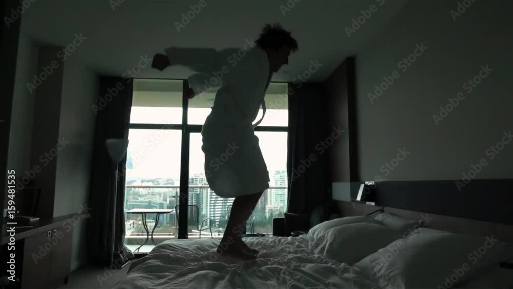 Slow motion. Happy man jumping on bed at hotel in bathrobe. Huge window. Cool apartments relaxation. Concept relax after hard working. Holiday. 240 fps