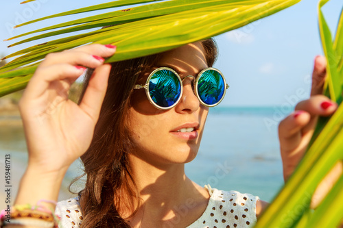 Close up portrait of a beautiful slim woman in fashion round sunglasses and white shirt push through palm leaves to the sun. Beauty sunshine cute girl on a sand beach with large stone. photo