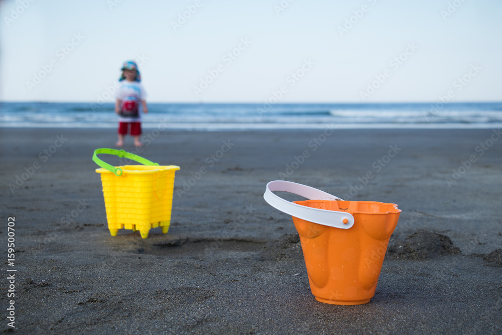 A pair of toy buckets at the beach with a child in the background A pair of toy buckets at the beach with a child in the background