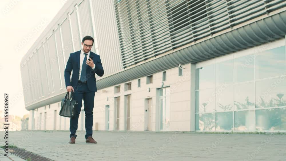 Stylish caucasian businessman talking on the cell phone, looking at the watch and running to a meeting near office building center. Man is late for work, meeting. Slow motion.