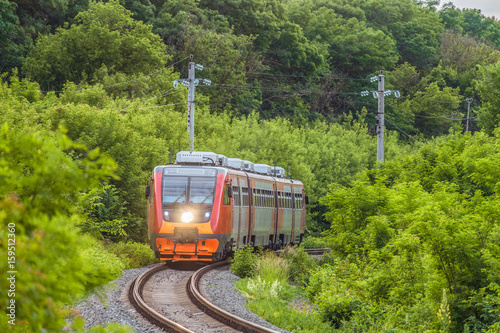 Modern commuter passenger red train is traveling along a single-track railway. Rolling stock with a searchlight on the turn of the railway among green trees.