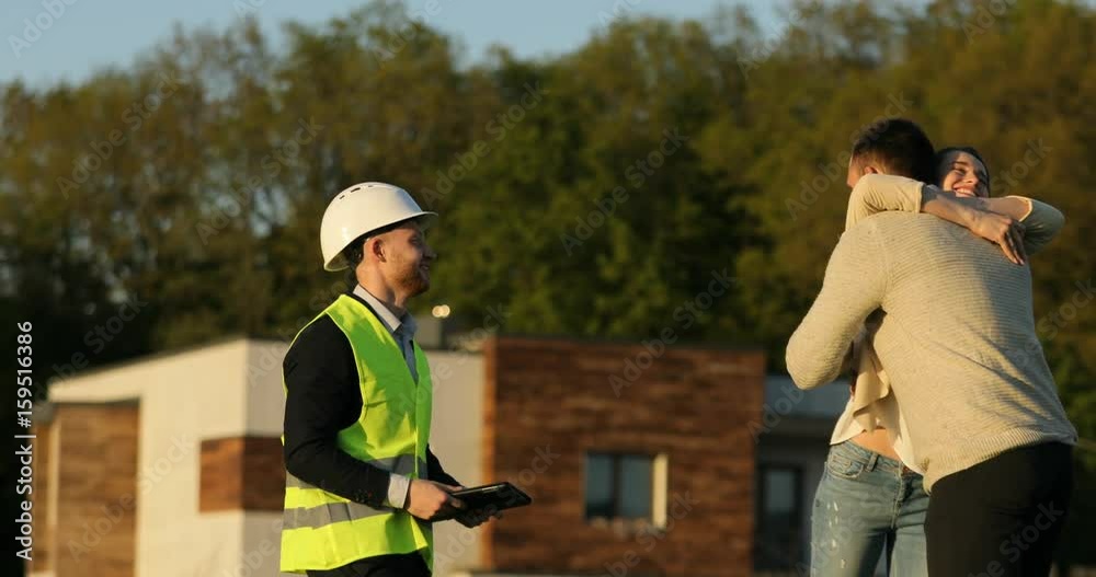 Caucasian architect in white helmet shaking hand of client and wife smiling and embracing her husband. Outdoor.