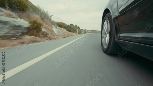 Wheels of the car are moving along the highway along the road markings. Car drives on asphalt level road in the middle of a mountainous area. View from on-board camera on weel and wing