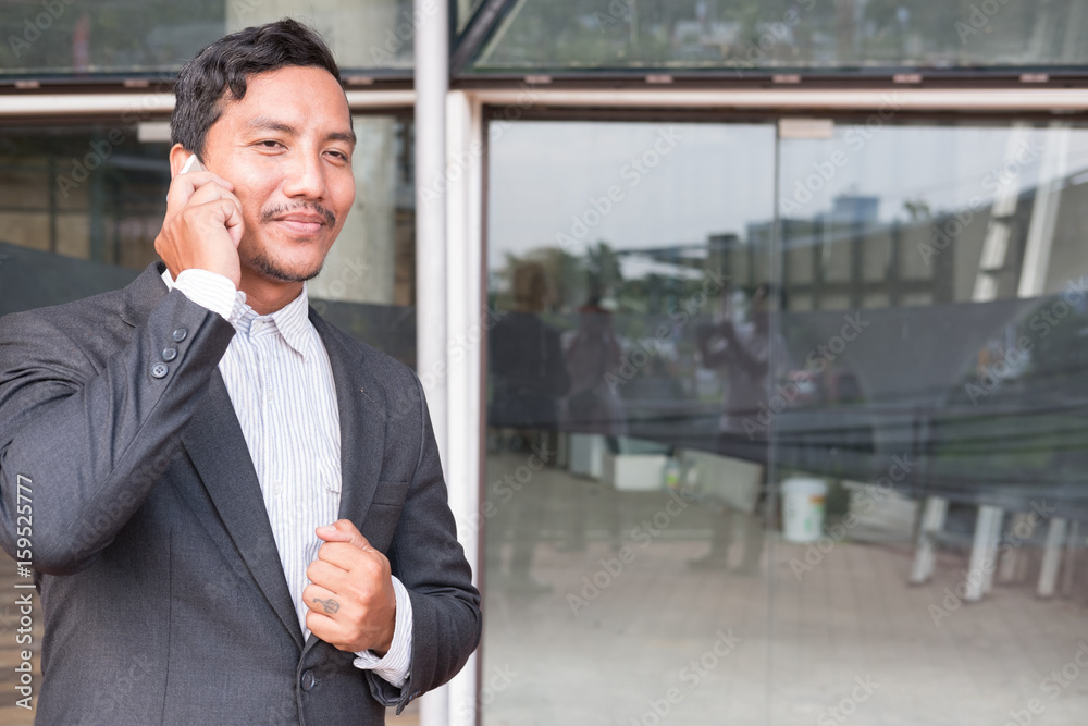 businessman in suit talking on smart phone while standing outside building. young asian male using cellphone for work at modern office corridor