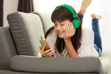 Beautiful Asian girl laying down on sofa and looking to smartphone with headphone on her head, concept for modern teenager free time
