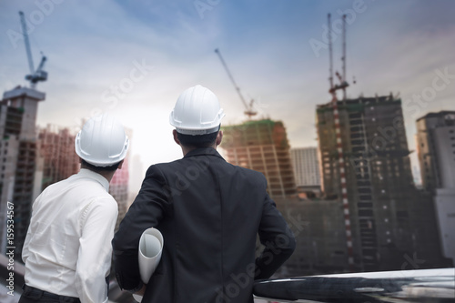 Asian businessman looking and point to finger away and engineer architect hold construction industrial plan background for working together as team concept, back view