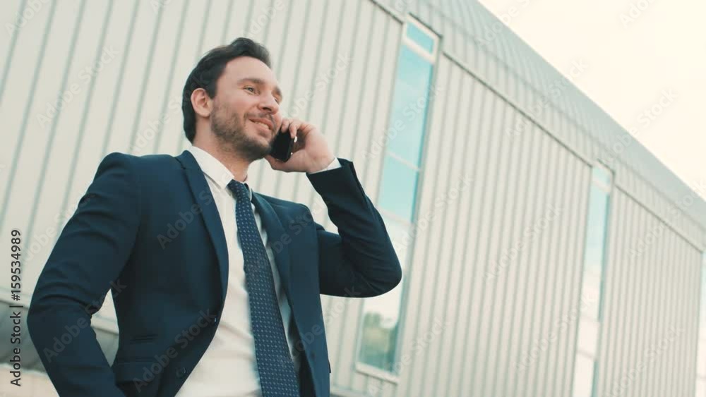 Attractive businessman talking on the mobile phone, feeling happy, hearing good news. Man raising hands. Outdoors in the city near office building. Close up Attractive businessman talking on the mobile phone, feeling happy, hearing good news. Man raising hands. Outdoors in the city near office building. Close up