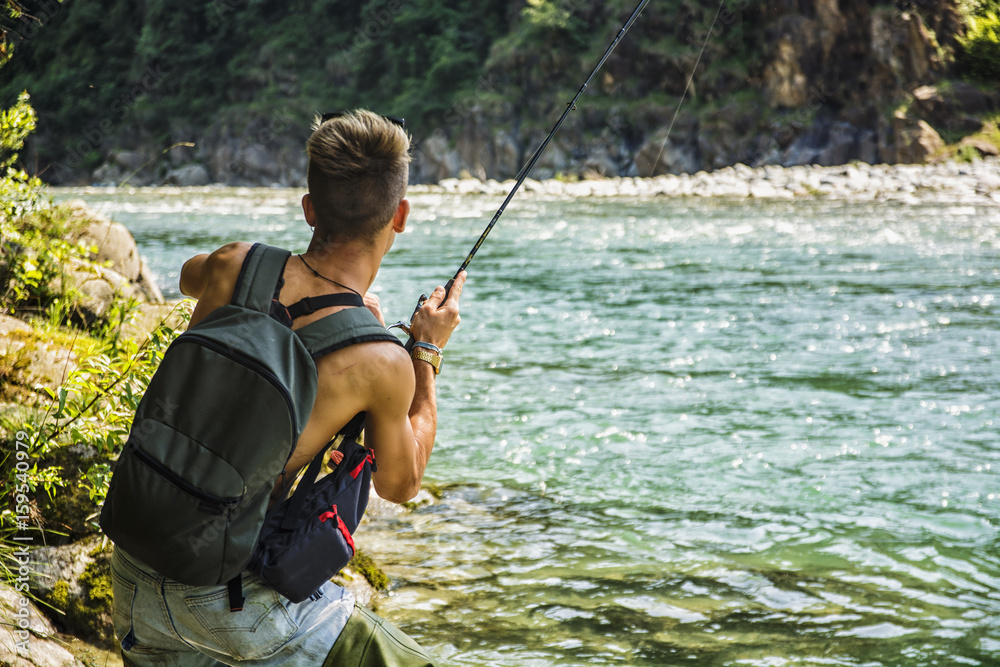 Young handsome male tourist standing and fishing in river in sunny day with rod Young handsome male tourist standing and fishing in river in sunny day with rod