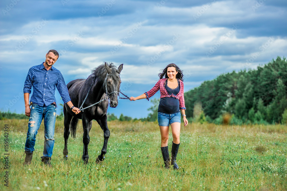 Awaiting the child, walking on the meadow. Young couple - she is a handsome brunette with long hair, pregnant; he is tall and brave, astride a black horse. Awaiting the child, walking on the meadow. Young couple - she is a handsome brunette with long hair, pregnant; he is tall and brave, astride a black horse.