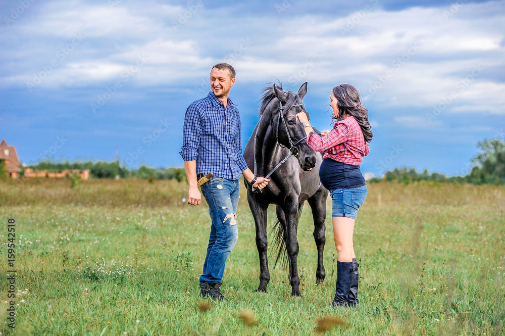 Awaiting the child, walking on the meadow. Young couple - she is a handsome brunette with long hair, pregnant; he is tall and brave, astride a black horse.