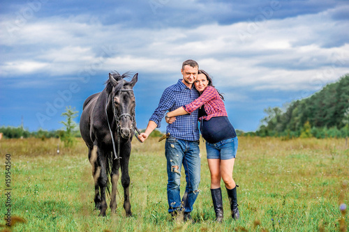 Awaiting the child, walking on the meadow. Young couple - she is a handsome brunette with long hair, pregnant; he is tall and brave, astride a black horse.