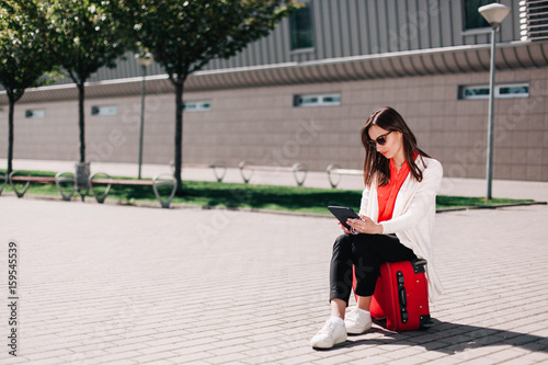 Woman reads something in the tablet sitting on red suitcase outside