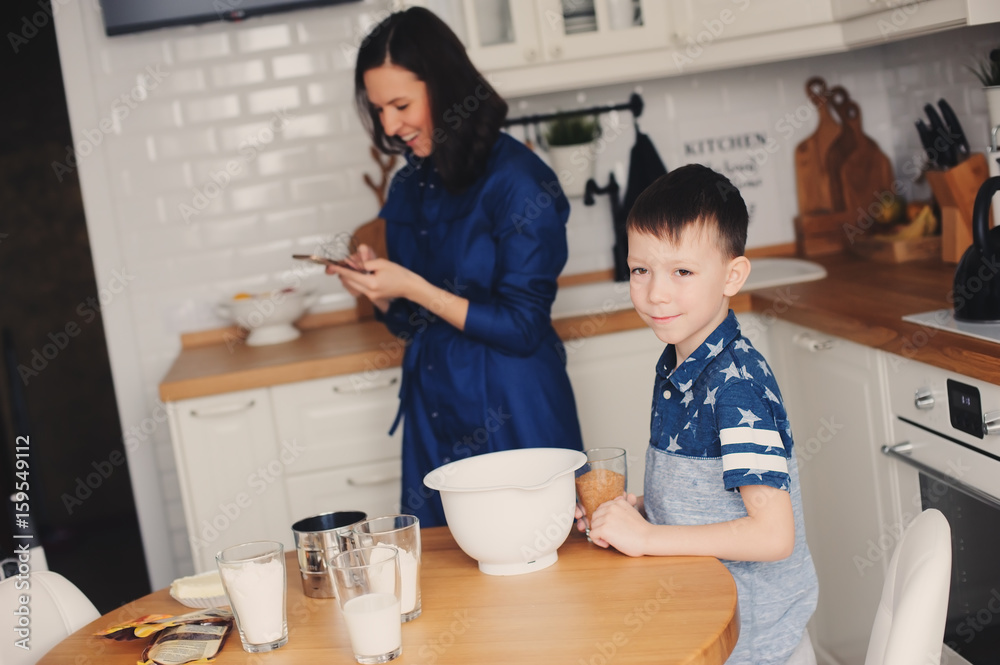 Kid boy helps mother to cook in modern white kitchen. Happy family in cozy weekend morning at home Kid boy helps mother to cook in modern white kitchen. Happy family in cozy weekend morning at home