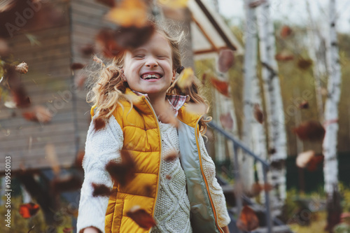 happy kid girl throwing leaves on the walk in autumn garden near wooden house  © mashiki