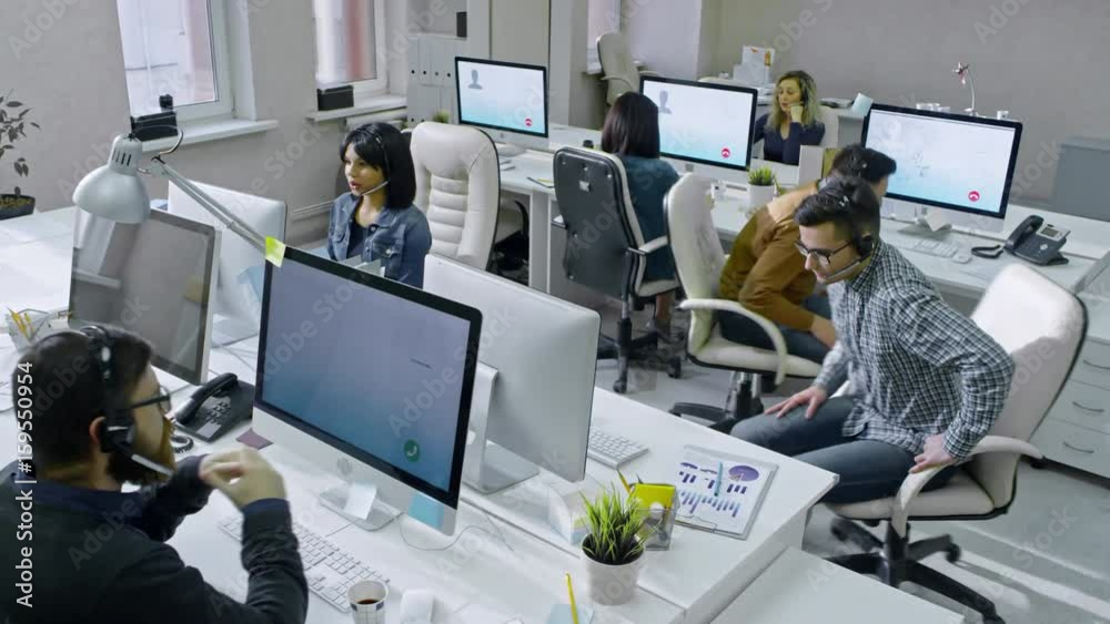 Crane shot of male workers of busy call centre sitting in office and discussing work, then returning to their computers and calling customers