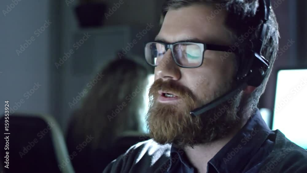 PAN of male call centre operator with beard wearing hands-free and talking on voice call with customer 