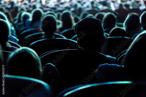 People, children, adults, parents in the theater watching the performance. People in the auditorium looking at the stage. Shooting from the back