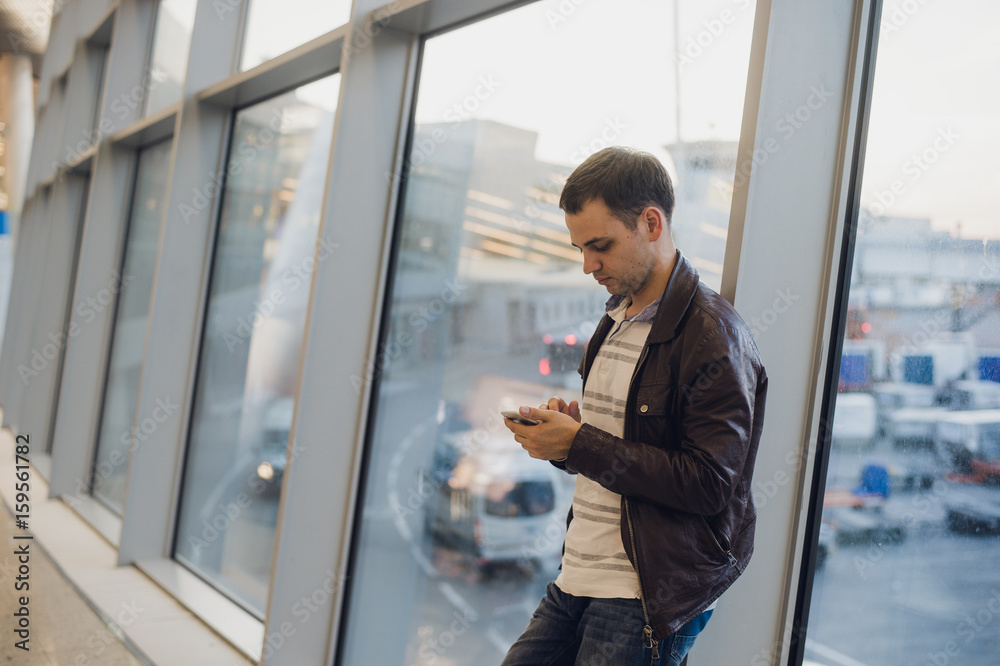 Portrait of young handsome person wearing casual style clothes standing near window in modern airport terminal. Traveler making call using smartphone.