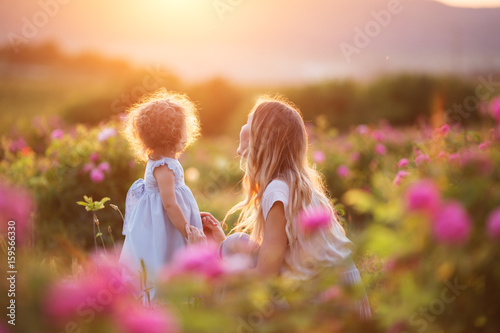 Beautiful child girl with young happy mother are wearing casual clothes walking in roses garden over sunset lights, summer time