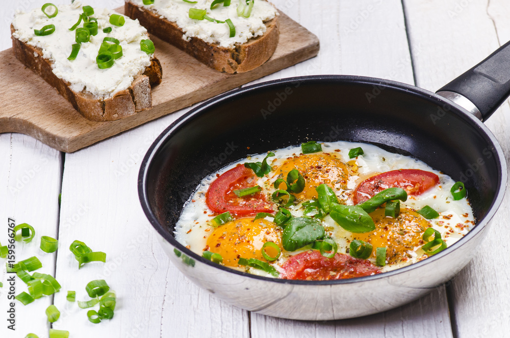 Pan of fried eggs with tomatoes, cheese, spring onion, herbs on a white table. Bread with spread. White wooden table. Concept of food. Breakfast time. Copy space. 