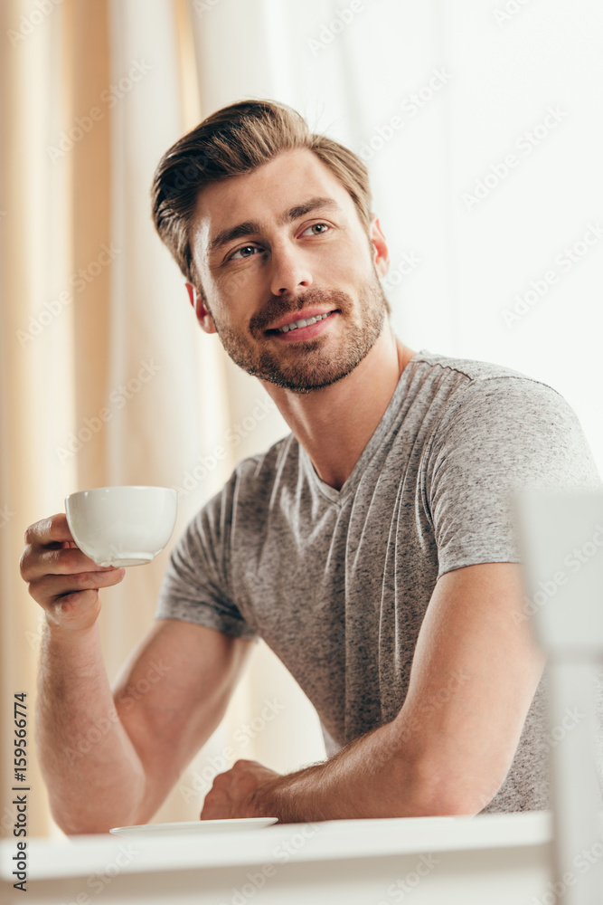 Smiling young man holding coffee cup and looking away at home Smiling young man holding coffee cup and looking away at home