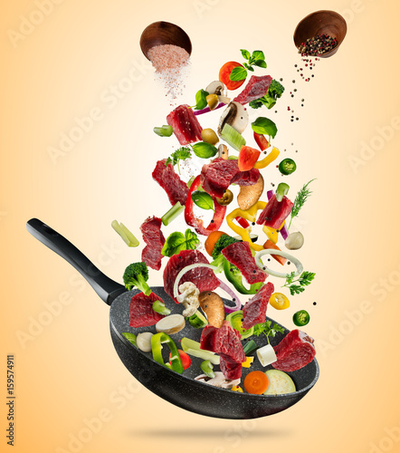 Fresh vegetables and beef meat flying into a pan on brown background