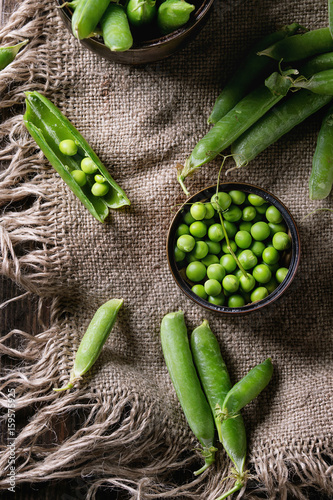 Young organic green pea pods and peas in can tin over old dark wooden planks with sackcloth textile background. Top view with space. Harvest, healthy eating.