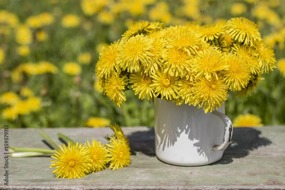 Fototapeta premium Bunch of beautiful yellow flowers of dandelion (taraxacum officinale) in an old enamel bowl stands on a wooden table against the background of meadows.