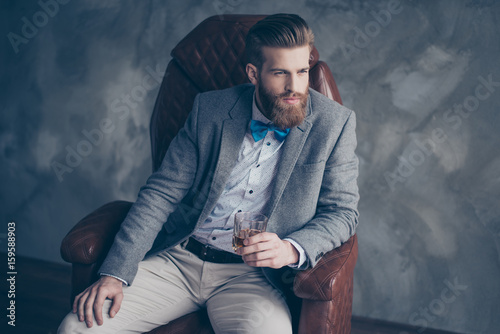 Successful young red bearded elegant businessman in suit with perfect hairstyle is drinking whiskey indoors, relaxing, sits on leather arm chair, looks harsh and virile