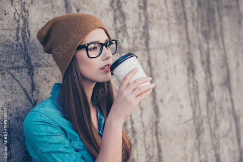 Charming dreamy young girl drinking tea near concrete wall outdoors. She is sleepy and relaxed, in casual jeans shirt, brown hat, black glasses