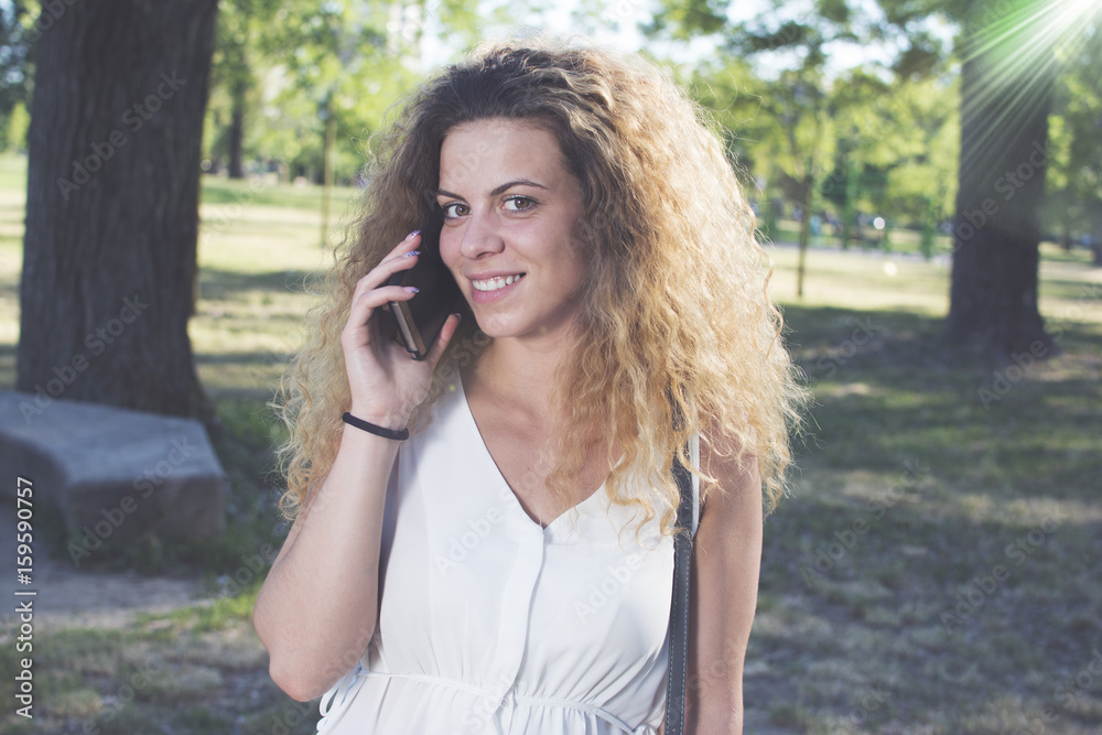 Girl calling on the phone and looking in the distance. Beautiful curly haired girl outdoors. Lens flare in the background. Girl calling on the phone and looking in the distance. Beautiful curly haired girl outdoors. Lens flare in the background.