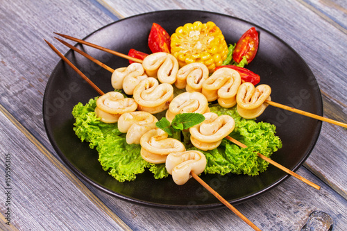 Grilled squid rings on skewers and vegetables on black plate