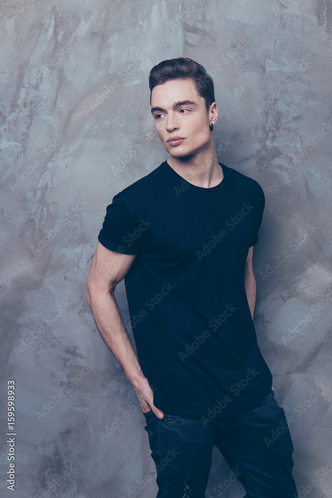 Handsome young male model is standing on the grey background. He is wearing black outfit, he put his hands in the back pockets of jeans