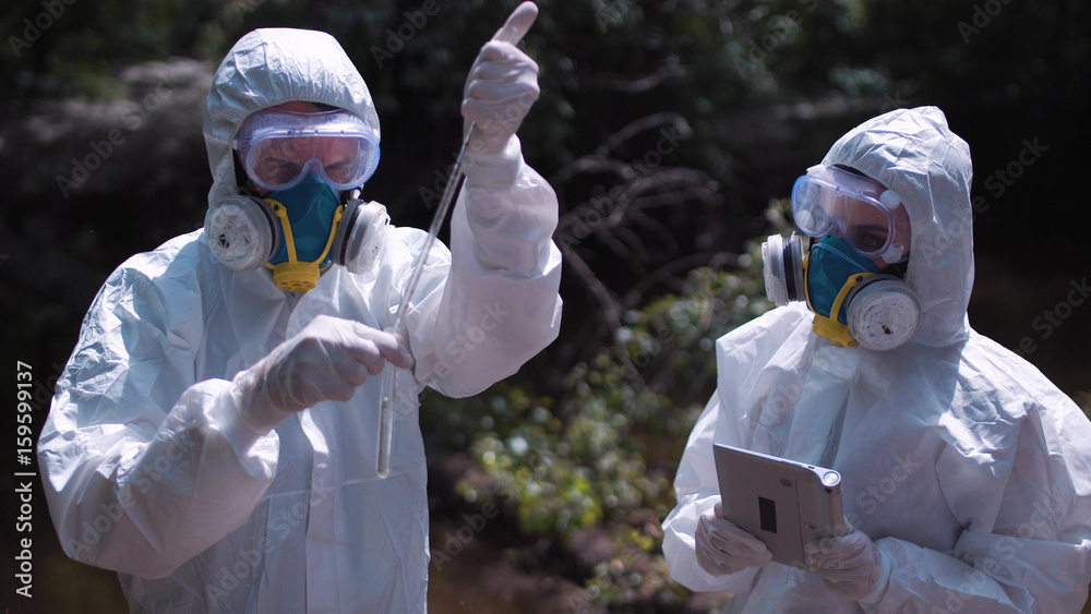 Φωτογραφία Two men in biohazard suits and masks sampling water from a stream or river pipetting a sample into a test tube for chemical analysis of pollutants