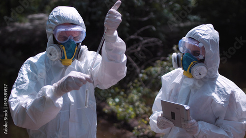 Two men in biohazard suits and masks sampling water from a stream or river pipetting a sample into a test tube for chemical analysis of pollutants.