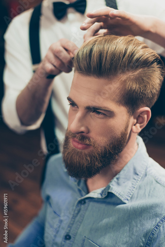 Close up cropped photo of a barber shop classy dressed stylist, who is doing a perfect hairstyle to a red bearded guy in caual jeans outfit