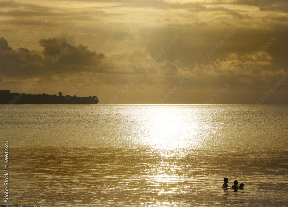 Obraz premium Small silhouettes of a man, woman and baby bathing in the ocean on a golden sunset with tropical land silhouette on the background